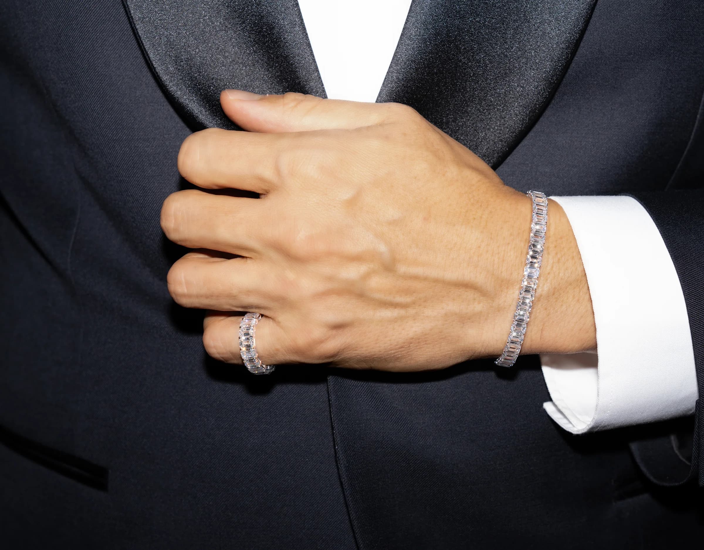A man's hand resting on a jacket lapel: a ring on the little finger, a bracelet at the cuff